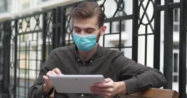 Tıbbi koruyucu maskeli yakışıklı genç adam bankta oturuyor ve tablet bilgisayarla çalışıyor. Sinema 4K 60fps eğik video