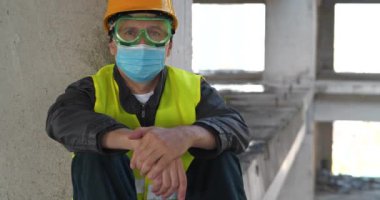 Tıbbi koruyucu yüz maskesi, gözlük ve başlık takan işçiler inşaat alanında oturur ve kameraya bakar. Sinema 4K 60fps eğik video