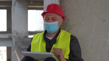 Mavi tıbbi koruyucu yüz maskesi ve kırmızı başlıklı mimar inşaat alanında oturup video bağlantısı üzerinden tablet bilgisayarla konuşuyor.