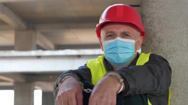 Koruyucu yüz maskeli, kırmızı başlıklı, kelepçeli müteahhit inşaat alanında oturur ve kameraya bakar.