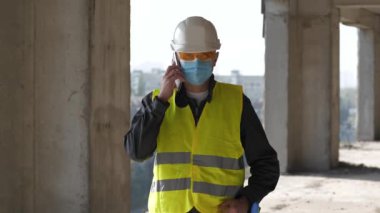 Beyaz kasklı ve tıbbi koruyucu maskeli işçiler inşaat alanında duruyor ve akıllı telefondan konuşuyor.
