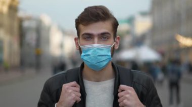 Tıbbi koruyucu maskeli, sırt çantalı genç adam sokakta durup kameraya bakıyor. 4K Geri Zaman Yavaş Hareket videosu