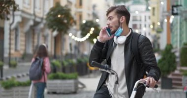 Sırt çantalı ve bisikletli Slow Motion Bisiklet kuryesi sokakta duruyor ve akıllı telefondan konuşuyor. Genç adam tıbbi koruyucu maskeni çıkar ve akıllı telefondan konuşmaya başla.