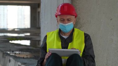 Kırmızı şapka ve tıbbi koruyucu maske takan misafir işçiler proje sahasının beton zemininde oturup tablet bilgisayarla çalışıyorlar.