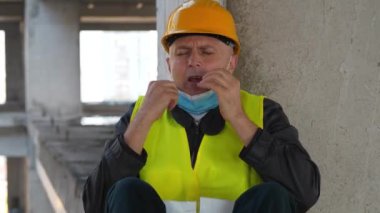 Kasklı ve tıbbi koruyucu maskeli işçiler inşaat alanının beton zemininde oturuyorlar. Kendini iyi hissetmiyor, hapşırıyor ve burnunu siliyor.