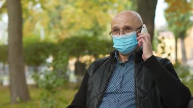 Tıbbi koruyucu maskeli kıdemli adam parkta bankta oturur, telefon numarasını çevirir ve akıllı telefondan konuşur. Covid konsept sağlık ve güvenlik, koronavirüs karantinası, sonraki dalga covid virüs koruması