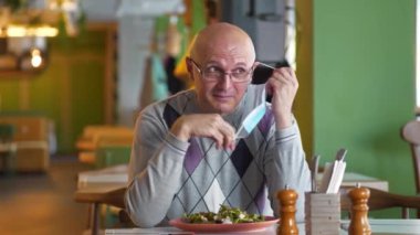 Kıdemli adam bir restoranda masaya oturur, akıllı telefonu masaya bırakır, tıbbi koruyucu maskeyi çıkarır, çatal ve bıçağı alır ve salata yemeye başlar.