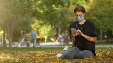 Slow Motion Young adlı tıbbi koruyucu maskeli kadın sonbahar parkında çimlerde oturur, akıllı telefon kullanır ve müzik dinler. Sonra maskesini çıkardı ve telefonsuz çalışmaya devam etti.