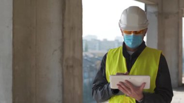 Beyaz başlıklı ve tıbbi koruyucu maskeli bir mimar inşaat alanında duruyor ve tablet bilgisayarla konuşuyor.