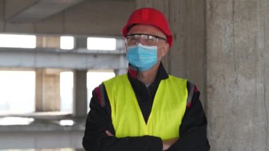 Tıbbi koruyucu maskeli, gözlüklü ve kırmızı başlıklı işçi inşaat alanında kameraya bakıyor. 4K 60fps panoramik video