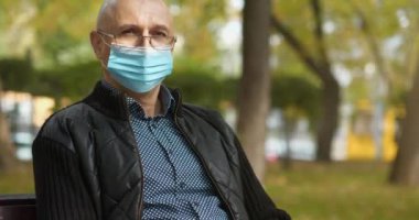 Gözlüklü ve tıbbi koruyucu maskeli kıdemli bir adam bankta oturur ve kameraya bakar. Sinema 4K 60fps eğik video