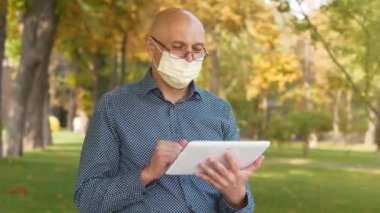 Tıbbi koruyucu maskeli kel adam şehir parkında oturuyor ve tablet bilgisayarla konuşuyor.