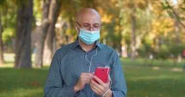 Tıbbi koruyucu maskeli kıdemli adam halka açık bir bahçede kürsüde oturuyor ve video bağlantısıyla iletişim kuruyor. Sinema 4K 60fps video
