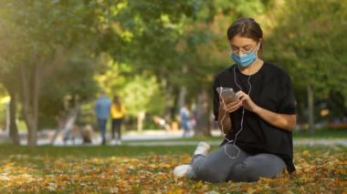 Medikal koruyucu maskeli genç bir kız sonbahar parkında çimenlerde oturup müzik dinler, sonra maskeyi çıkarır ve o olmadan akıllı telefonu kullanmaya devam eder.