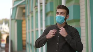 Slow Motion Genç yakışıklı tıbbi koruyucu maskeli ve siyah gömlekli adam hastane binasının yanında duruyor.