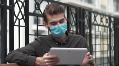Ağır çekim maskeli genç adam kürsüye oturur ve tablet bilgisayar kullanır, fotoğraf çeker ve video kaydeder.