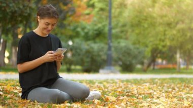Slow Motion Genç güzel kız sonbahar şehir parkında çimenlerde oturuyor, akıllı telefonundaki fotoğraflara bakıyor, gülümsüyor, gülüyor ve keyfi yerinde.