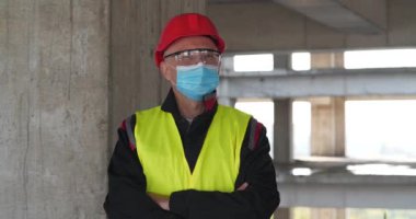 Tıbbi koruyucu maskeli, gözlüklü ve kırmızı başlıklı bir işçi proje alanında kameraya bakıyor. Sinema 4K 60fps panoramik video