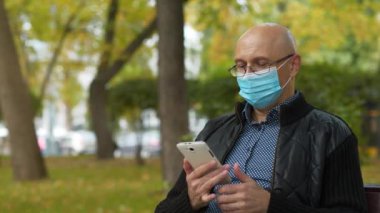 Koruyucu maskeli kel, orta yaşlı bir adam parkta bankta oturur, telefon numarasını çevirir ve akıllı telefondan konuşur. Covid konsept sağlık ve güvenlik, koronavirüs karantinası, sonraki dalga covid virüs koruması