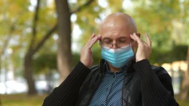 Gözlüklü ve tıbbi koruyucu maskeli hasta kıdemli adam şehir parkındaki bankta oturuyor, kendini iyi hissetmiyor ve başı ağrıyor, kafasına masaj yapıyor.