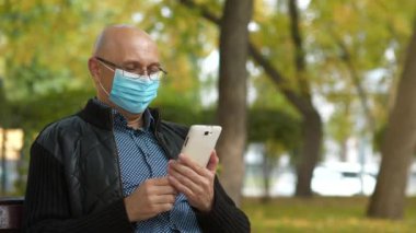 Slow Motion 'ın koruyucu maskeli kıdemli adamı parkta bankta oturuyor ve akıllı telefon aracılığıyla iletişim kuruyor. Covid konsept sağlık ve güvenlik, N1H1 koronavirüs karantinası, sosyal uzaklık