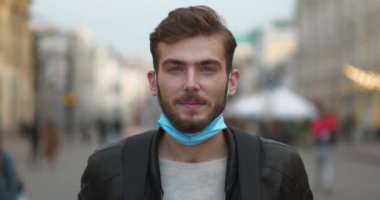 Çenesinde tıbbi koruyucu maskesi olan genç adam sokakta dikilip kameraya bakıyor. Sinema 4K Ters Zaman Yavaş Hareket videosu