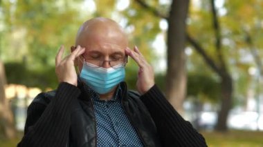 Şehir parkında gözlüklü ve mavi koruyucu maskeli hasta kıdemli adam kendini iyi hissetmiyor ve başı ağrıyor, kafasına masaj yapıyor.