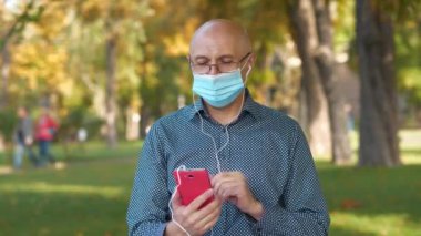 Tıbbi koruyucu maskeli kıdemli adam parkta bankta oturuyor ve akıllı telefon aracılığıyla iletişim kuruyor. Covid konsept sağlık ve güvenlik, koronavirüs karantinası, sosyal uzaklık, bir sonraki dalga covid virüs koruması