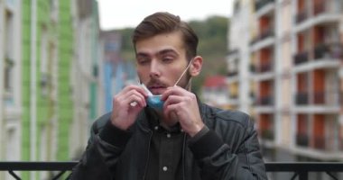 Genç hasta adam tıbbi koruyucu maskesini çıkarıyor, hapşırıyor, burnunu siliyor ve ceketinin fermuarını çekiyor. Sinema 4K 60fps video