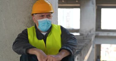 Sarı kasklı ve tıbbi koruyucu maskeli işçiler inşaat alanında oturuyorlar. Öksürüyor ve iyi hissetmiyor. Sinema 4K 60fps video