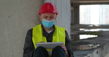 Kırmızı başlıklı ve tıbbi koruyucu maskeli işçiler inşaat alanında oturup tablet bilgisayarla çalışıyorlar. Sinema 4K 60fps panoramik video