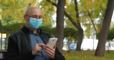 Tıbbi koruyucu maskeli kıdemli adam bankta oturuyor ve akıllı telefonunu kullanıyor. Covid konsept sağlık ve güvenlik, koronavirüs karantinası, bir sonraki dalga covid virüs koruması. Sinema 4K 60fps video