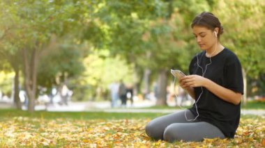 Genç ve güzel bir kız sonbahar parkında çimlerde oturur, akıllı telefon kullanır ve müzik dinler.