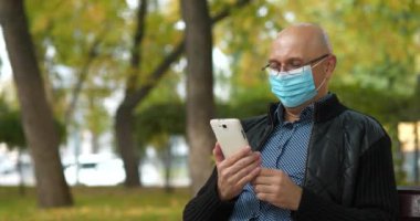 Tıbbi koruyucu maskeli kel bir adam parkta bankta oturuyor ve akıllı telefon aracılığıyla iletişim kuruyor. Covid konsept sağlık ve güvenlik, N1H1 koronavirüs karantinası, sosyal mesafe. Sinema 4K 60fps video