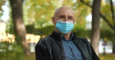 Mavi tıbbi koruyucu maskeli ve gözlüklü kıdemli adam şehir parkında bir bankta oturur ve kameraya bakar. Arka plandaki arabalar yolda gidiyor. Sinema 4K 60fps panoramik video