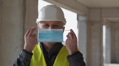 Slow Motion Workman inşaat alanında beyaz başlık takıyor tıbbi koruyucu maske takıyor ve kameraya bakıyor.