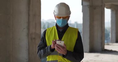 Kasketli ve koruyucu maskeli bir işçi proje alanında duruyor ve verileri akıllı telefonuna yüklüyor. Sinema 4K 60fps panoramik video