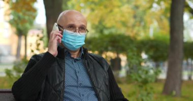 Tıbbi koruyucu maskeli kıdemli adam parkta bankta oturuyor ve akıllı telefondan konuşuyor. Covid konsept sağlık ve güvenlik, koronavirüs karantinası, bir sonraki dalga covid virüs koruması. Sinema 4K 60fps video