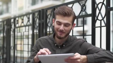 Slow Motion genç adam bankta oturuyor ve tablet bilgisayarındaki fotoğraflara bakıyor, gülümsüyor ve keyfi yerinde.