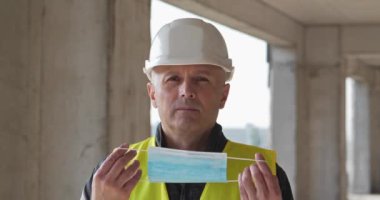 Proje alanındaki beyaz başlıklı adam mavi tıbbi koruyucu maske takıyor ve kameraya bakıyor. Sinema 4K 60fps video