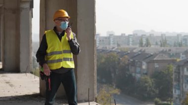 Kasklı, gözlüklü ve tıbbi koruyucu maskeli işçiler proje alanında duruyor ve taşınabilir radyo vericisiyle konuşuyor.