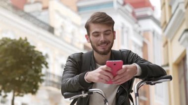Sırt çantalı bisikletli kurye akıllı telefonundaki fotoğraflara bakıyor. Bisikletli genç adam sokakta duruyor, akıllı telefon kullanıyor ve gülümsüyor.