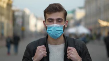 Sırt çantalı tıbbi koruyucu maskeli bir öğrenci sokakta dikiliyor ve kameraya bakıyor.