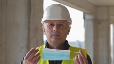 İnşaat alanındaki beyaz başlıklı işçi tıbbi koruyucu maske takıyor ve kameraya bakıyor.