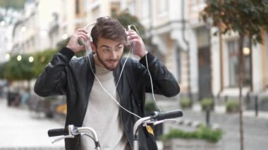 Bisikletli kurye sokakta duruyor, kulaklık takıyor ve akıllı telefonuyla müzik dinliyor.