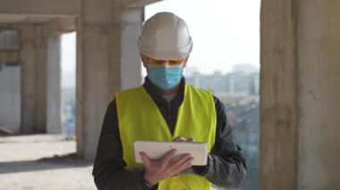 Başlık ve tıbbi maske takan işçiler inşaat alanında duruyor ve verileri tablet bilgisayara giriyor.