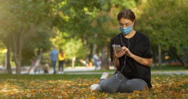 Güz parkında çimenlikte oturan güzel bir kız akıllı telefonundaki fotoğraflara bakar. Sonra maskesini çıkardı ve maskesiz fotoğraflara bakmaya devam etti. Sinema 4K 60fps video