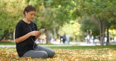 Güz parkında çimlere oturup müzik dinleyen güzel bir kız. Sinema 4K 60fps video