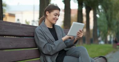 Genç bir kadın sonbahar şehir meydanında bankta oturuyor ve tablet bilgisayardaki fotoğraflara bakıyor. Sinema 4K 60fps panoramik video