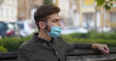 Genç adam şehir parkındaki bankta oturur, mavi tıbbi koruyucu maskeyi ayarlar ve etrafına bakar. Sinema 4K 60fps panoramik video